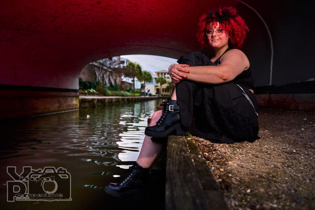 That Witchy Friend, with curly red hair and glasses, sits on the edge of a canal under a bridge, illuminated by a red glow. They wear a black gothic-style outfit with chain accessories and black boots, with their arms wrapped around their knees. The canal water reflects the surrounding structures and sky.