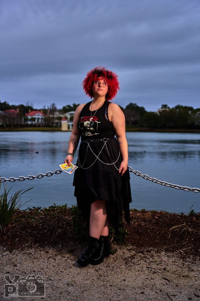 A female with curly red hair and glasses stands near a body of water, holding a tarot card in one hand. They wear a black gothic-style outfit with chain accessories, a black tank top with a skull design, and black boots. The background features houses, trees, and a cloudy sky.
